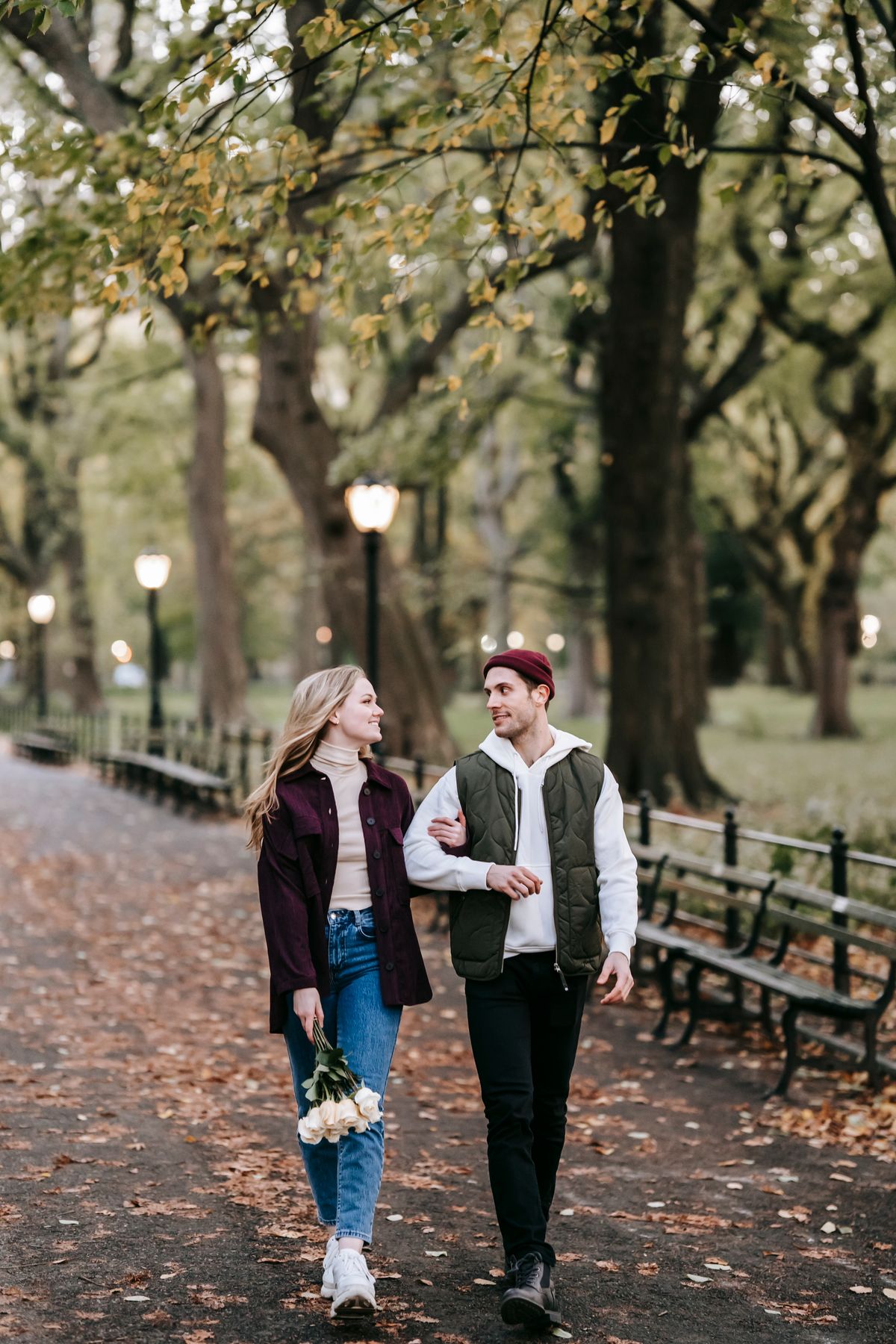 A young couple strolling through a park amid falling autumn leaves, holding flowers