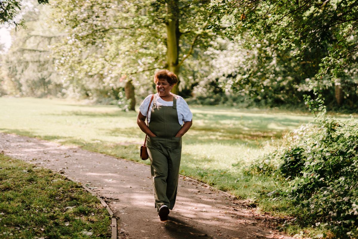 A content woman walks a sunlit, tree-lined park path with no phone in sight