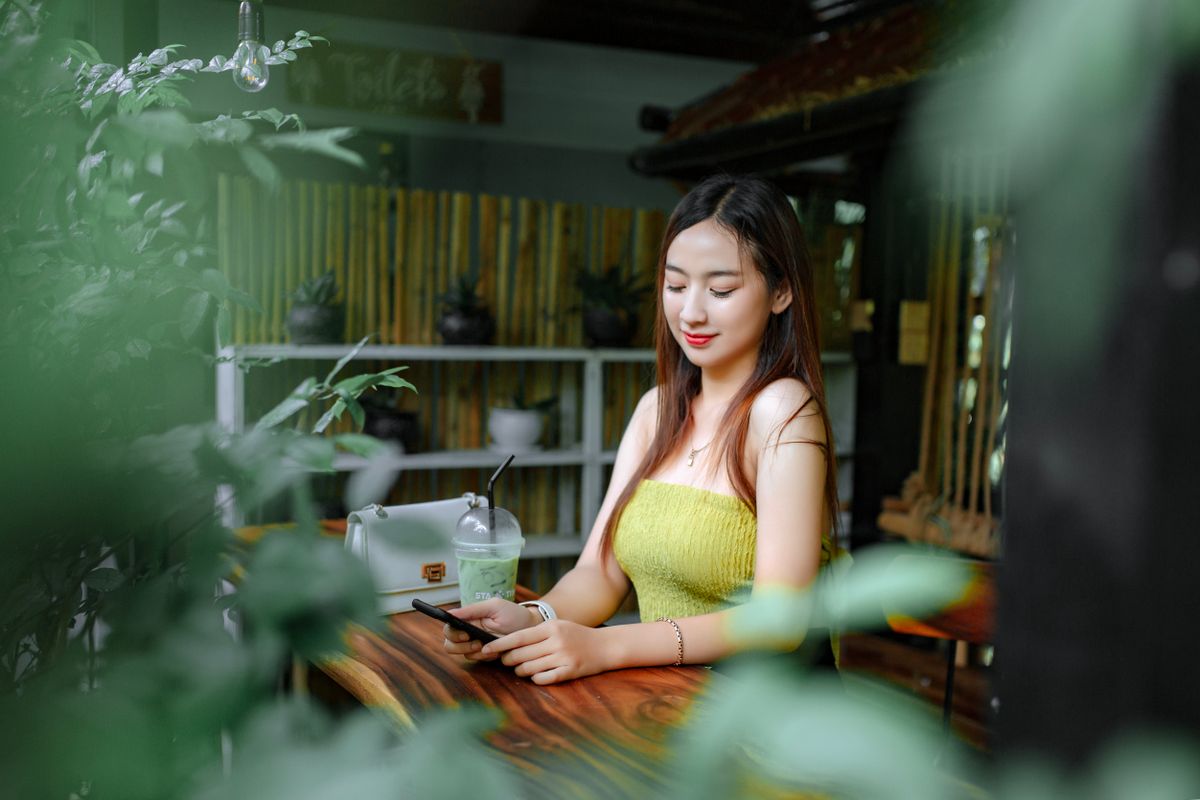 A young woman smiles softly at her phone in a plant-filled cafe, looking calm and hopeful